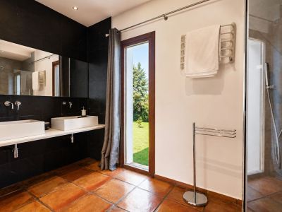 Salle de bain spacieuse avec carrelage en terre cuite, double vasque rectangulaire blanche sur un mur carrelé noir. Grande fenêtre offrant une vue sur le jardin et douche vitrée.