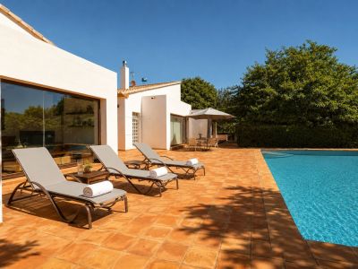 Vaste terrasse carrelée en terre cuite avec piscine, trois chaises longues et un espace repas ombragé. La façade blanche est agrémentée de grandes fenêtres reflétant la végétation luxuriante.