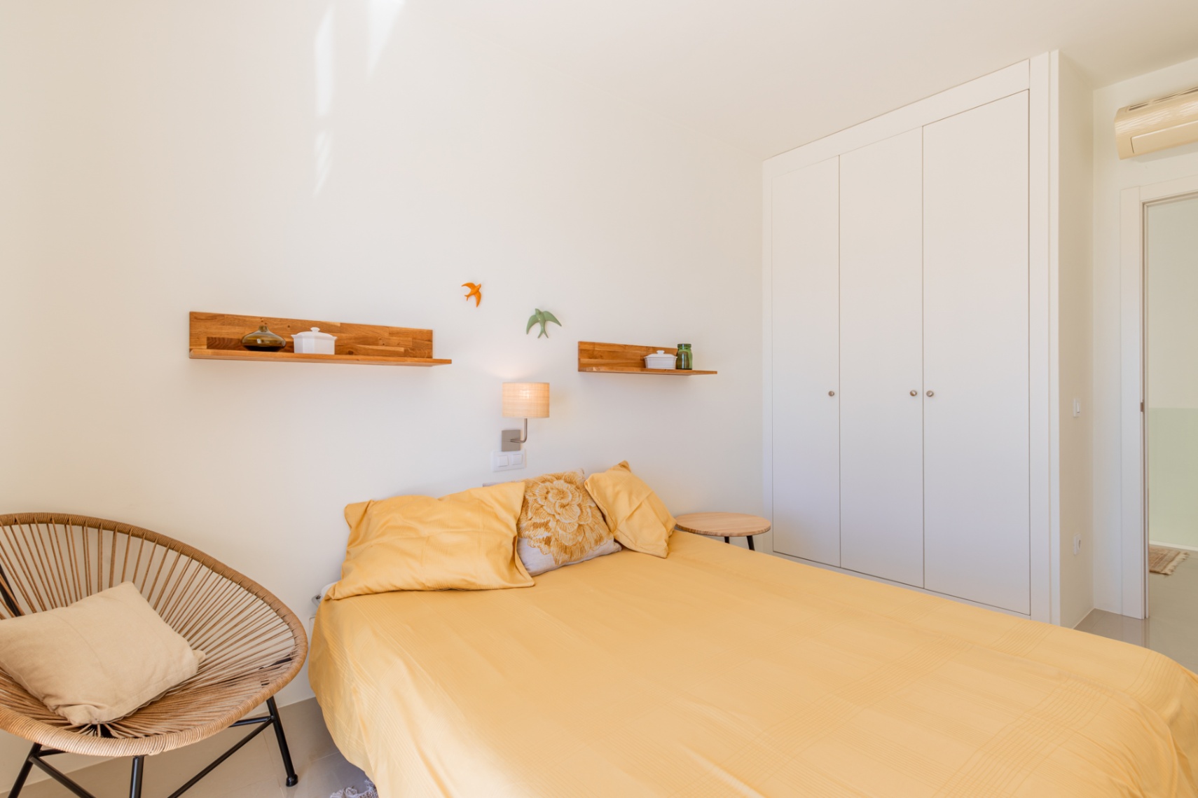 Chambre lumineuse dotée d'un lit avec literie jaune, d'une armoire encastrée blanche et d'un fauteuil en osier. Des étagères en bois et une applique murale complètent le décor. Chambre lumineuse dotée d'un lit avec literie jaune, d'une armoire encastrée blanche et d'un fauteuil en osier. Des étagères en bois et une applique murale complètent le décor.