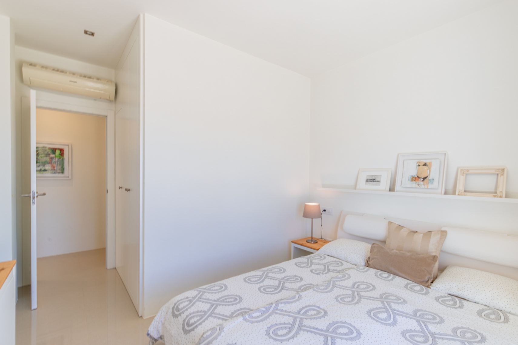 Chambre moderne aux murs blancs, carrelage clair et lit avec couette à motifs. Comprend une table de chevet en bois, une étagère décorative, des armoires intégrées et la climatisation. Chambre moderne aux murs blancs, carrelage clair et lit avec couette à motifs. Comprend une table de chevet en bois, une étagère décorative, des armoires intégrées et la climatisation.