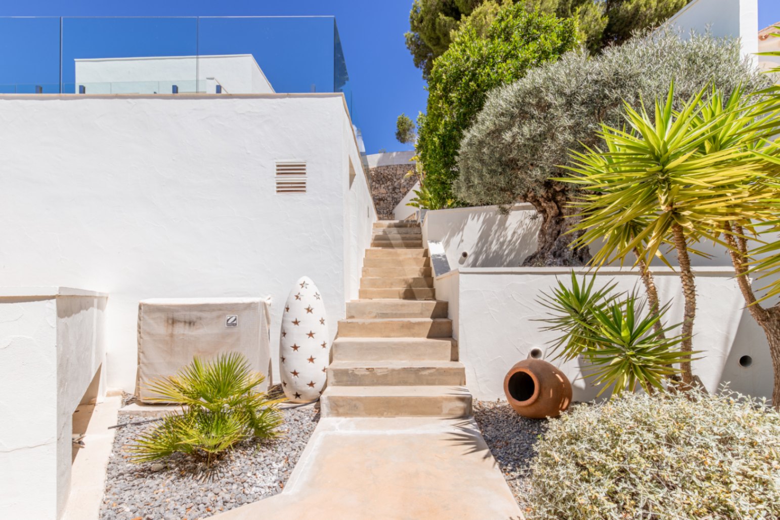 Escalier extérieur avec murs en stuc blanc, plantes méditerranéennes et aménagement paysager de gravier, menant à un niveau supérieur sous un ciel bleu. Escalier extérieur avec murs en stuc blanc, plantes méditerranéennes et aménagement paysager de gravier, menant à un niveau supérieur sous un ciel bleu.