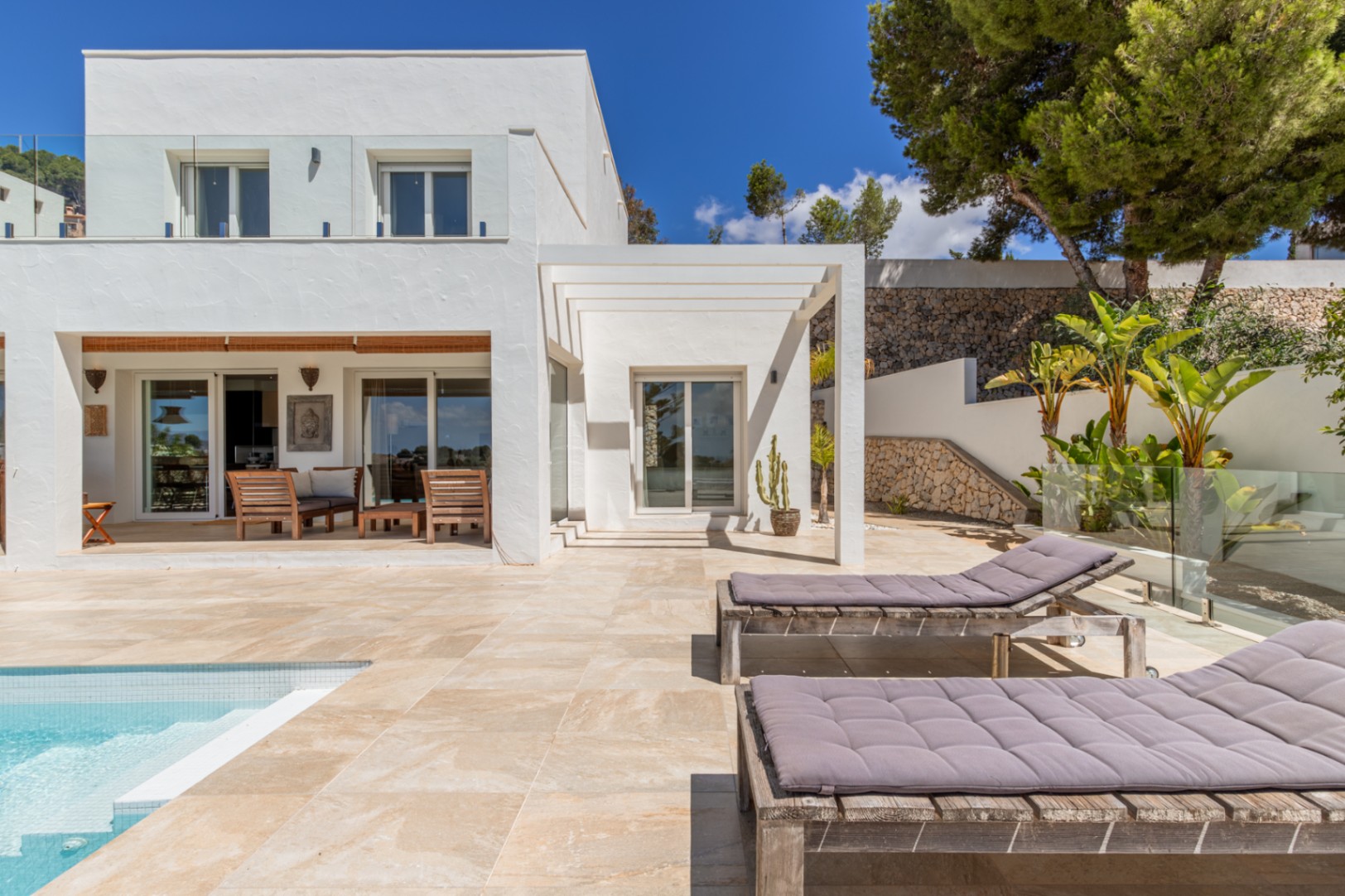 Extérieur de villa blanche moderne avec piscine carrelée, terrasse ensoleillée dotée de chaises longues en bois et un espace de vie couvert avec mobilier en bois, encadré par un jardin en terrasses avec murs en pierre. Extérieur de villa blanche moderne avec piscine carrelée, terrasse ensoleillée dotée de chaises longues en bois et un espace de vie couvert avec mobilier en bois, encadré par un jardin en terrasses avec murs en pierre.
