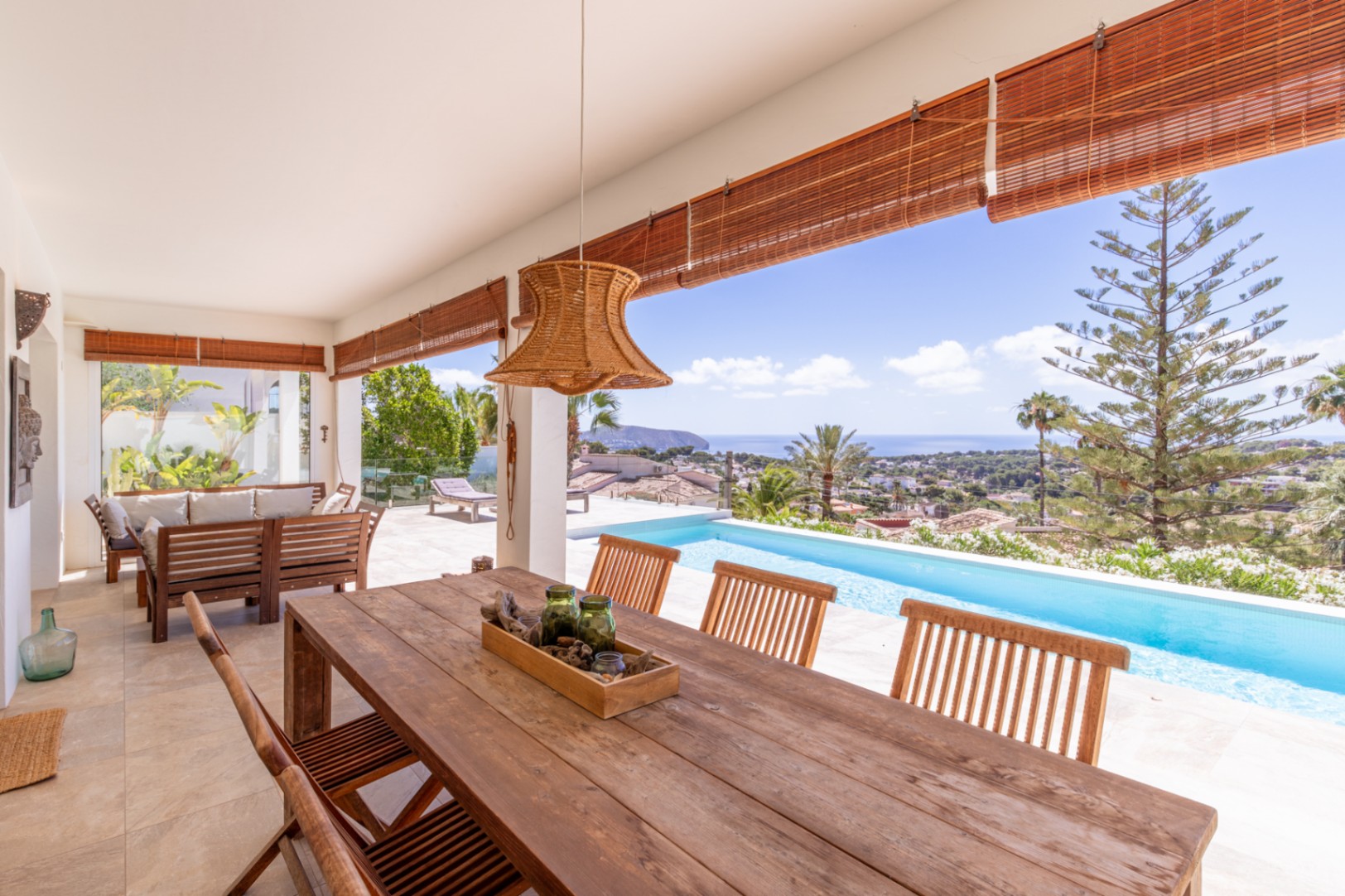Grande terrasse couverte avec table à manger en bois, suspension en rotin et vue imprenable sur la piscine à débordement et la mer Méditerranée. Grande terrasse couverte avec table à manger en bois, suspension en rotin et vue imprenable sur la piscine à débordement et la mer Méditerranée.