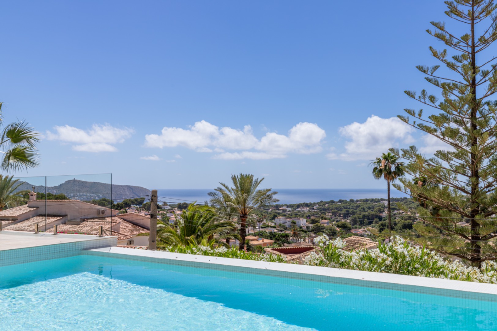 Piscine carrelée bleue avec vue panoramique sur la Méditerranée, toits traditionnels et végétation luxuriante sous un ciel clair. Garde-corps en verre. Piscine carrelée bleue avec vue panoramique sur la Méditerranée, toits traditionnels et végétation luxuriante sous un ciel clair. Garde-corps en verre.