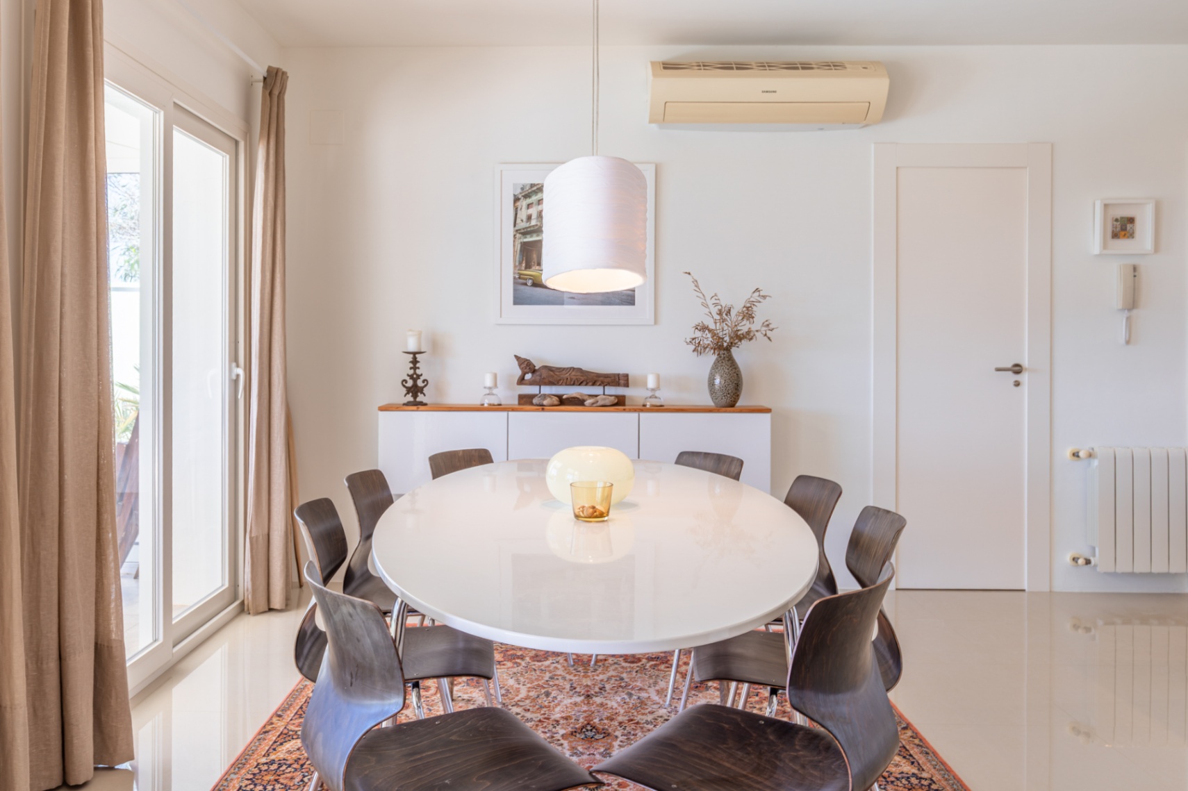 Salle à manger lumineuse avec table ovale blanche et chaises en bois foncé sur tapis. Porte coulissante vitrée avec rideaux et buffet décoratif. Salle à manger lumineuse avec table ovale blanche et chaises en bois foncé sur tapis. Porte coulissante vitrée avec rideaux et buffet décoratif.