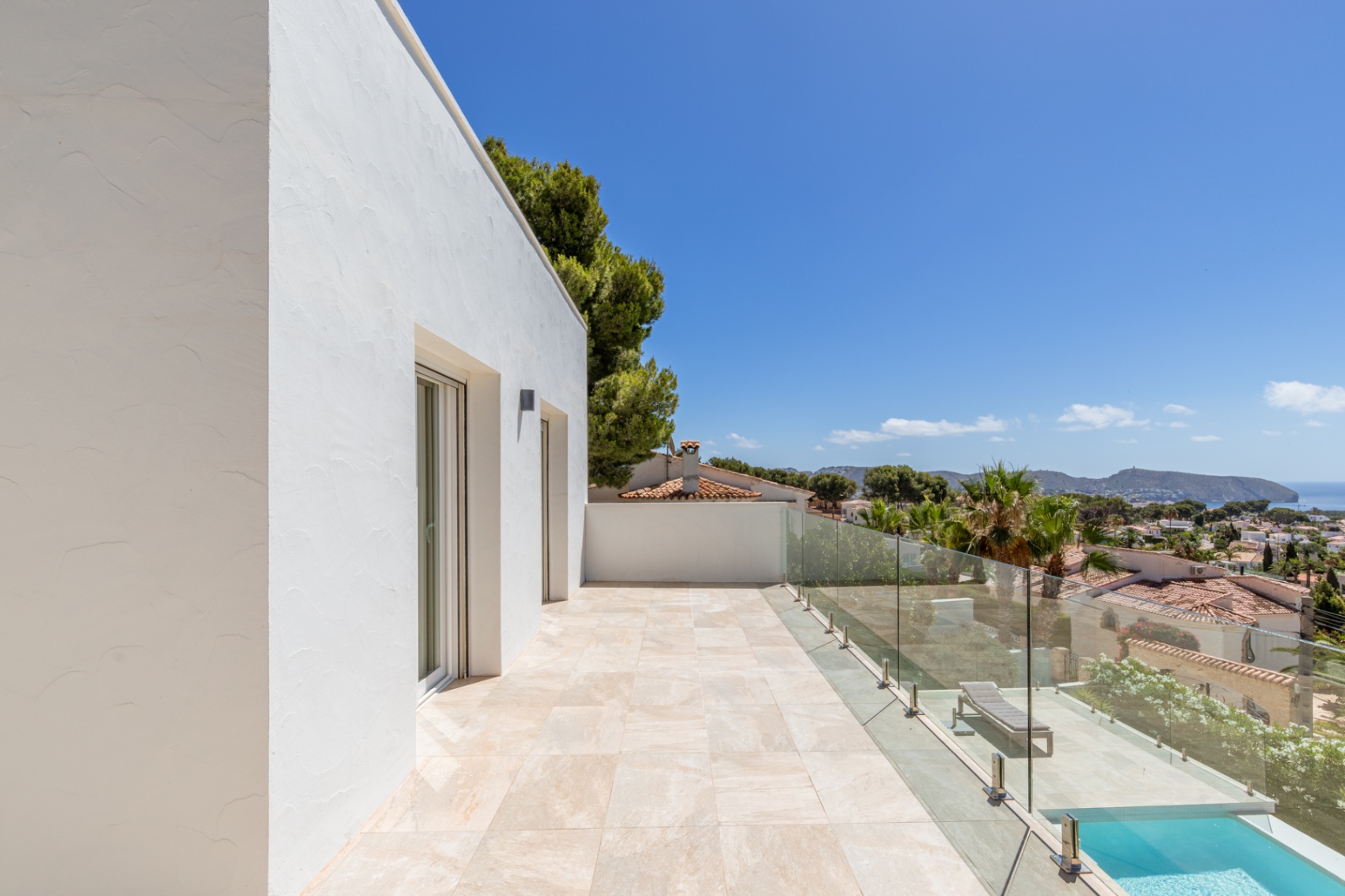Terrasse supérieure avec carrelage clair et balustrade en verre, offrant une vue panoramique sur la Méditerranée et le paysage côtier. Des portes vitrées et une piscine inférieure sont visibles. Terrasse supérieure avec carrelage clair et balustrade en verre, offrant une vue panoramique sur la Méditerranée et le paysage côtier. Des portes vitrées et une piscine inférieure sont visibles.