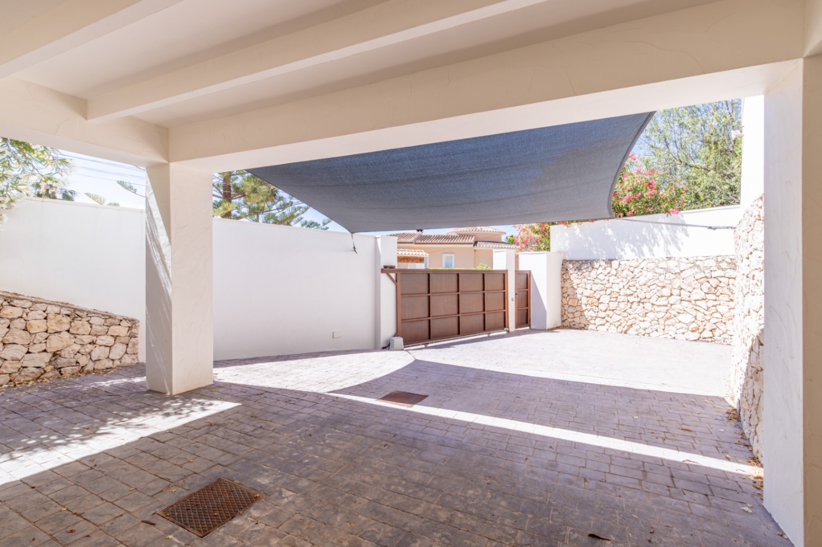 Vaste carport privé avec pavé texturé, voile d'ombrage protecteur et portail coulissant métallique sécurisé, encadré de murs blancs et en pierre naturelle. Vaste carport privé avec pavé texturé, voile d'ombrage protecteur et portail coulissant métallique sécurisé, encadré de murs blancs et en pierre naturelle.