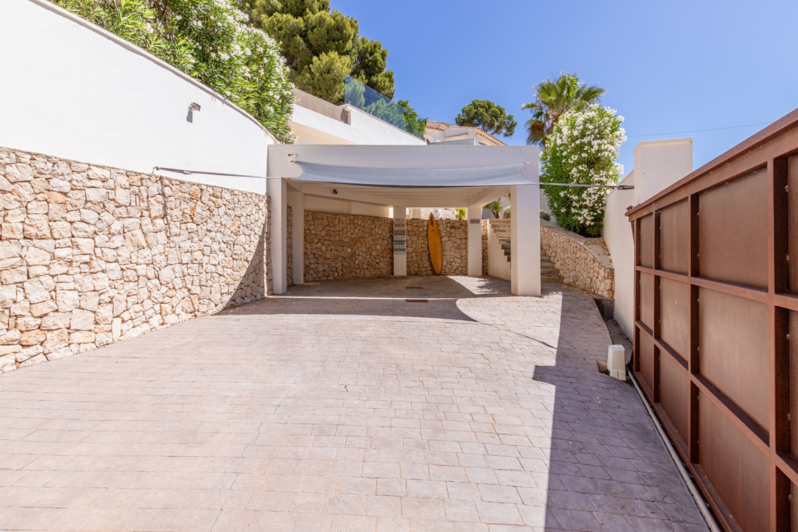 Vaste parking extérieur avec carport et revêtement pavé. On y voit des murs en pierre naturelle, une végétation méditerranéenne luxuriante et un portail métallique. Vaste parking extérieur avec carport et revêtement pavé. On y voit des murs en pierre naturelle, une végétation méditerranéenne luxuriante et un portail métallique.