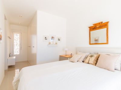 Chambre lumineuse avec lit blanc, tête de lit capitonnée et table de chevet en bois. Un miroir orné et un couloir avec fenêtre étroite donnant sur un mur de pierre.