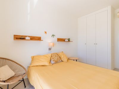 Chambre lumineuse dotée d'un lit avec literie jaune, d'une armoire encastrée blanche et d'un fauteuil en osier. Des étagères en bois et une applique murale complètent le décor.