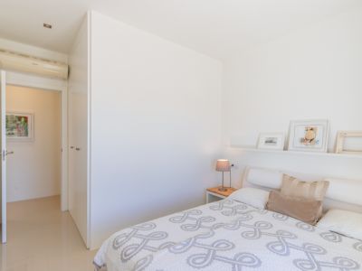 Chambre moderne aux murs blancs, carrelage clair et lit avec couette à motifs. Comprend une table de chevet en bois, une étagère décorative, des armoires intégrées et la climatisation.