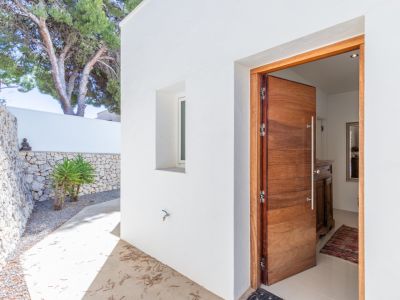 Entrée principale avec une porte moderne en bois massif, encadrée par des murs blancs et un mur en pierres sèches. Un chemin en béton mène au hall d'entrée lumineux.
