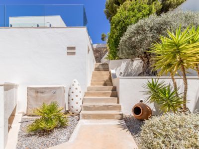 Escalier extérieur avec murs en stuc blanc, plantes méditerranéennes et aménagement paysager de gravier, menant à un niveau supérieur sous un ciel bleu.