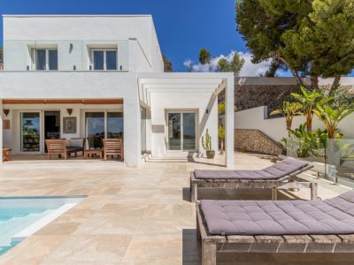 Extérieur de villa blanche moderne avec piscine carrelée, terrasse ensoleillée dotée de chaises longues en bois et un espace de vie couvert avec mobilier en bois, encadré par un jardin en terrasses avec murs en pierre.