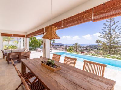 Grande terrasse couverte avec table à manger en bois, suspension en rotin et vue imprenable sur la piscine à débordement et la mer Méditerranée.