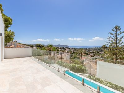 Grande terrasse extérieure carrelée de couleur claire avec balustrade en verre. Vues panoramiques sur la Méditerranée, la baie et le paysage urbain, avec piscine étroite et végétation.