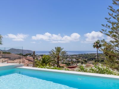Piscine carrelée bleue avec vue panoramique sur la Méditerranée, toits traditionnels et végétation luxuriante sous un ciel clair. Garde-corps en verre.