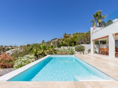 Piscine rectangulaire aux eaux bleues claires avec bordure épurée, entourée d'une terrasse carrelée et d'une végétation méditerranéenne luxuriante. Une salle à manger extérieure couverte est visible.
