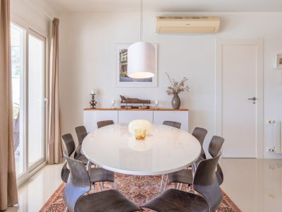 Salle à manger lumineuse avec table ovale blanche et chaises en bois foncé sur tapis. Porte coulissante vitrée avec rideaux et buffet décoratif.