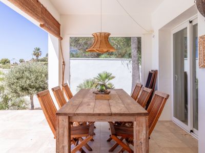 Terrasse couverte avec table et chaises en bois rustique, éclairée par une suspension tressée. Vues directes sur la verdure méditerranéenne luxuriante.