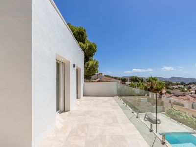 Terrasse supérieure avec carrelage clair et balustrade en verre, offrant une vue panoramique sur la Méditerranée et le paysage côtier. Des portes vitrées et une piscine inférieure sont visibles.
