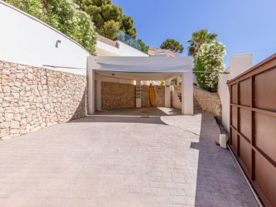 Vaste parking extérieur avec carport et revêtement pavé. On y voit des murs en pierre naturelle, une végétation méditerranéenne luxuriante et un portail métallique.