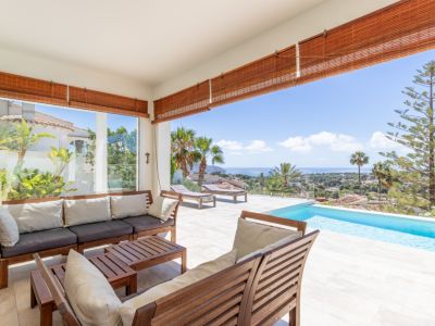 Vaste terrasse couverte avec mobilier de salon en bois, carrelage clair et piscine à débordement, offrant une vue panoramique sur la Méditerranée.