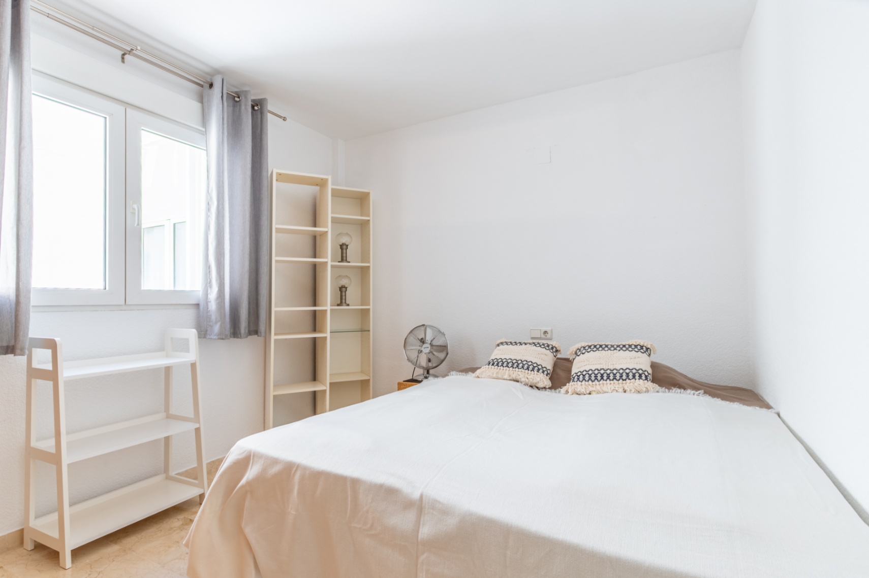 Chambre lumineuse avec lit double, étagères en bois clair et ventilateur. La fenêtre avec rideaux gris apporte une lumière naturelle abondante à l'espace aux murs blancs et au sol en marbre.