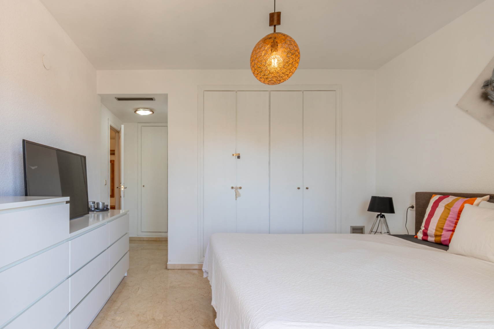 Chambre principale avec placards intégrés blancs, sol en marbre clair et luminaire suspendu sphérique. Comprend une commode avec TV et accès au couloir.