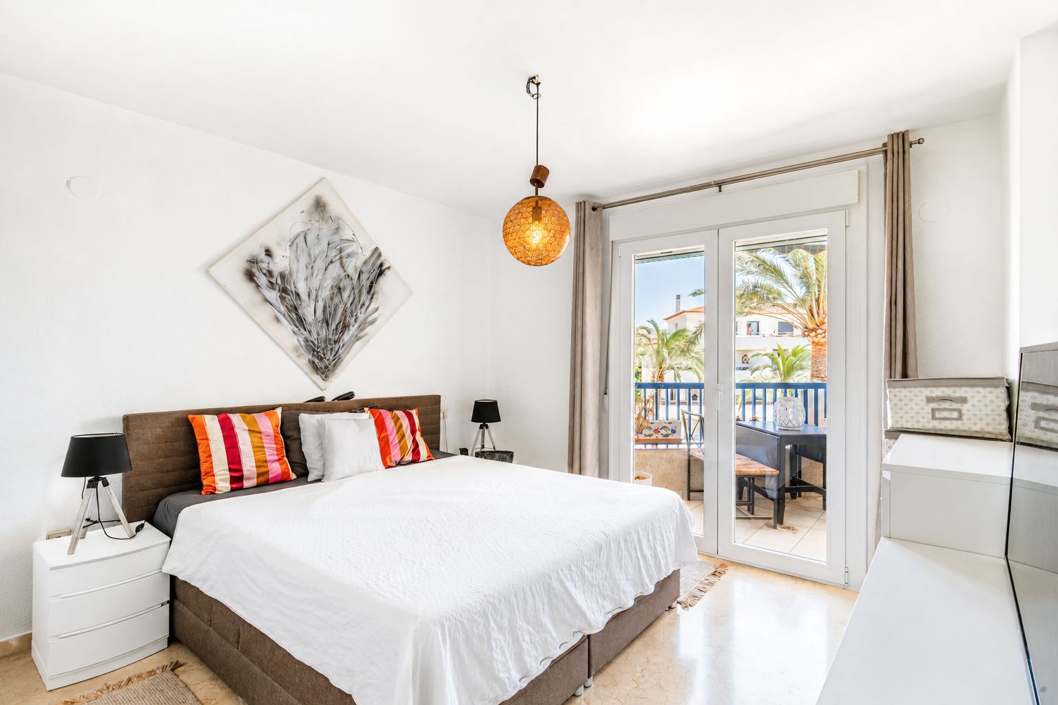 Chambre principale lumineuse avec accès direct à une terrasse privée, sol en marbre et vue sur les palmiers. Une lampe suspendue distinctive et de l'art abstrait complètent l'espace.