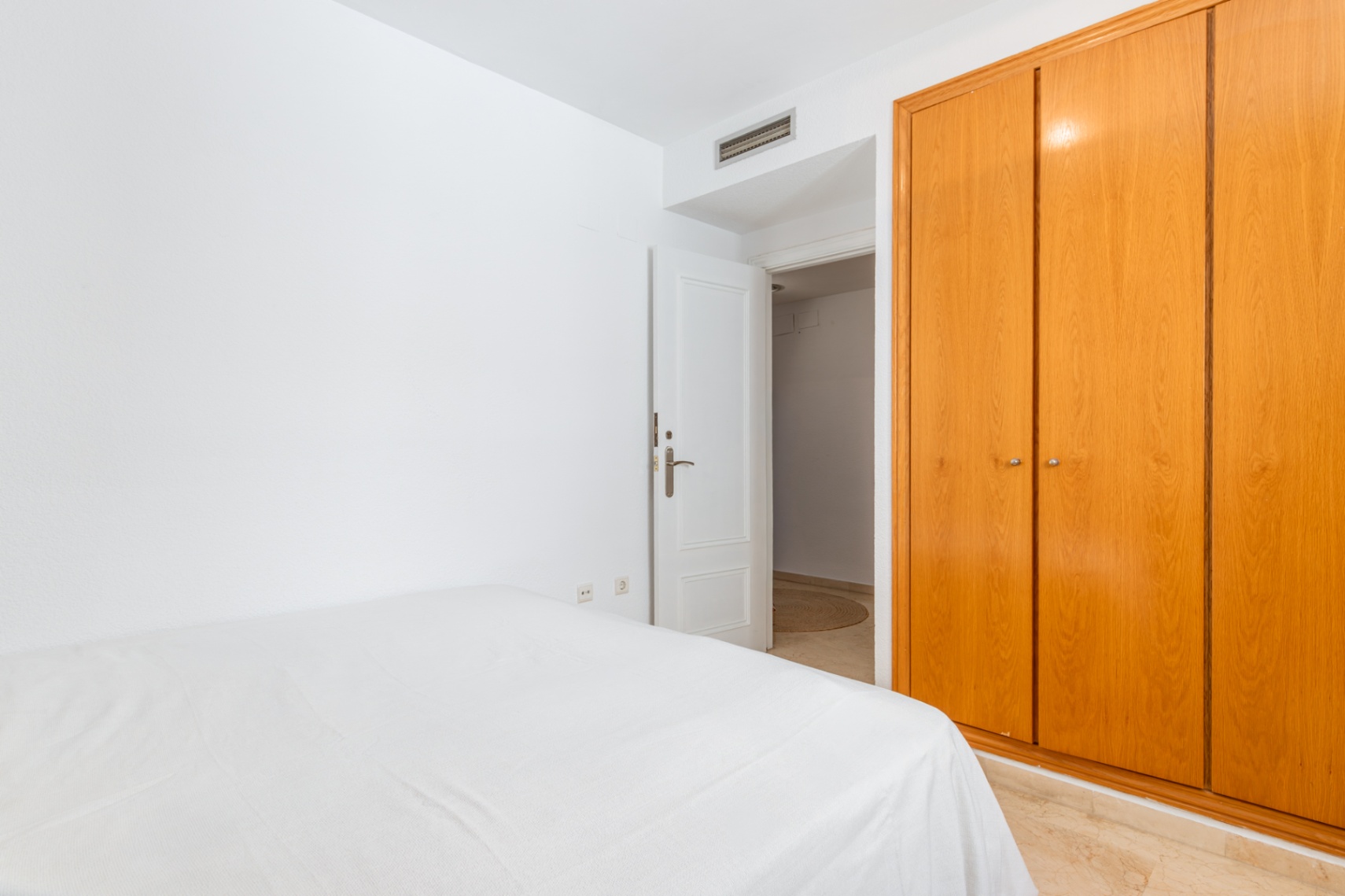 Chambre secondaire avec grand placard intégré en bois clair et lit habillé de linge blanc. Le sol présente un carrelage effet marbre et une porte ouverte mène à une autre pièce.