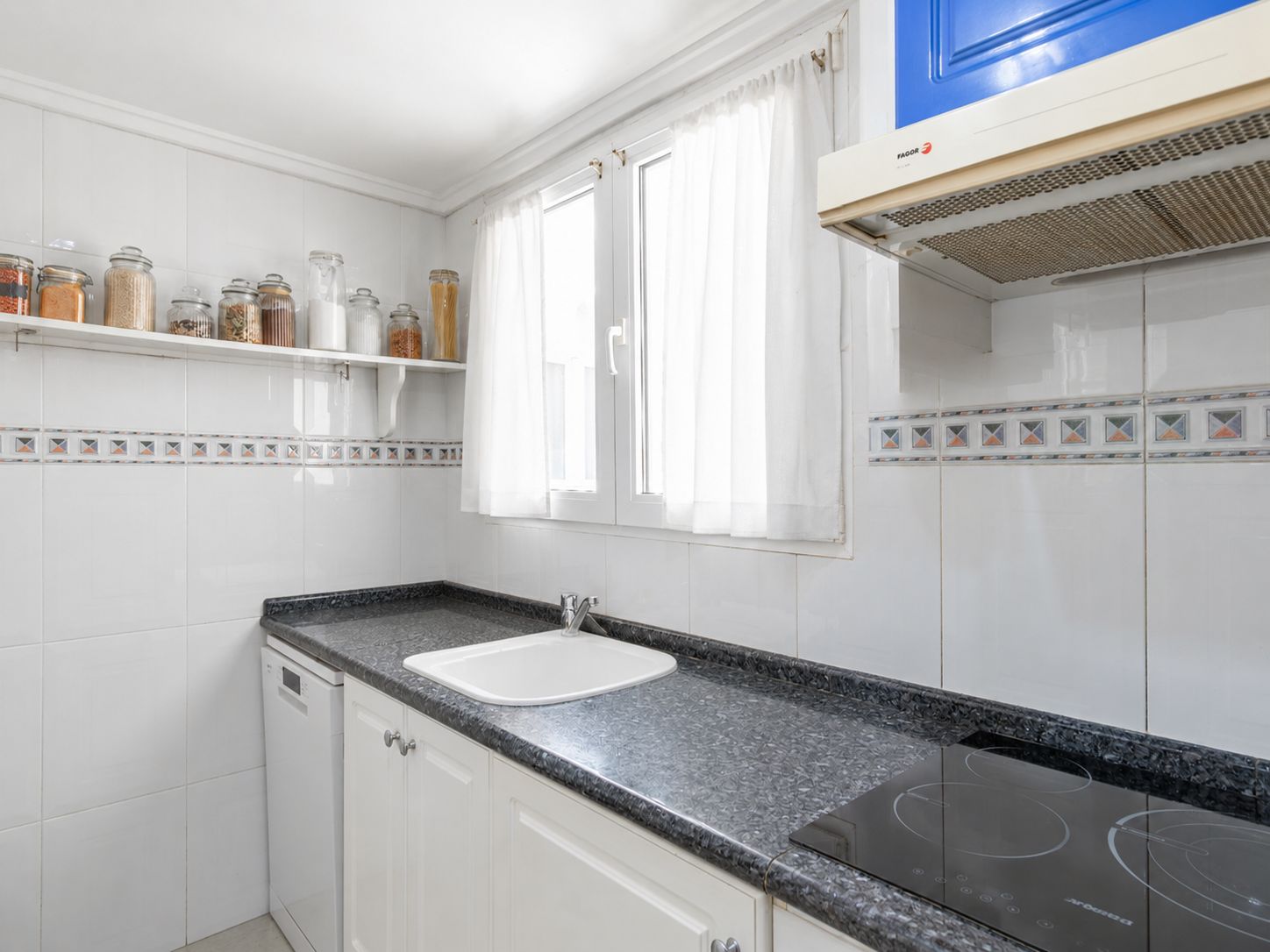 Cuisine indépendante avec plan de travail en granit foncé, évier blanc et plaque à induction. Murs carrelés blancs avec frise décorative et fenêtre apportant lumière naturelle.