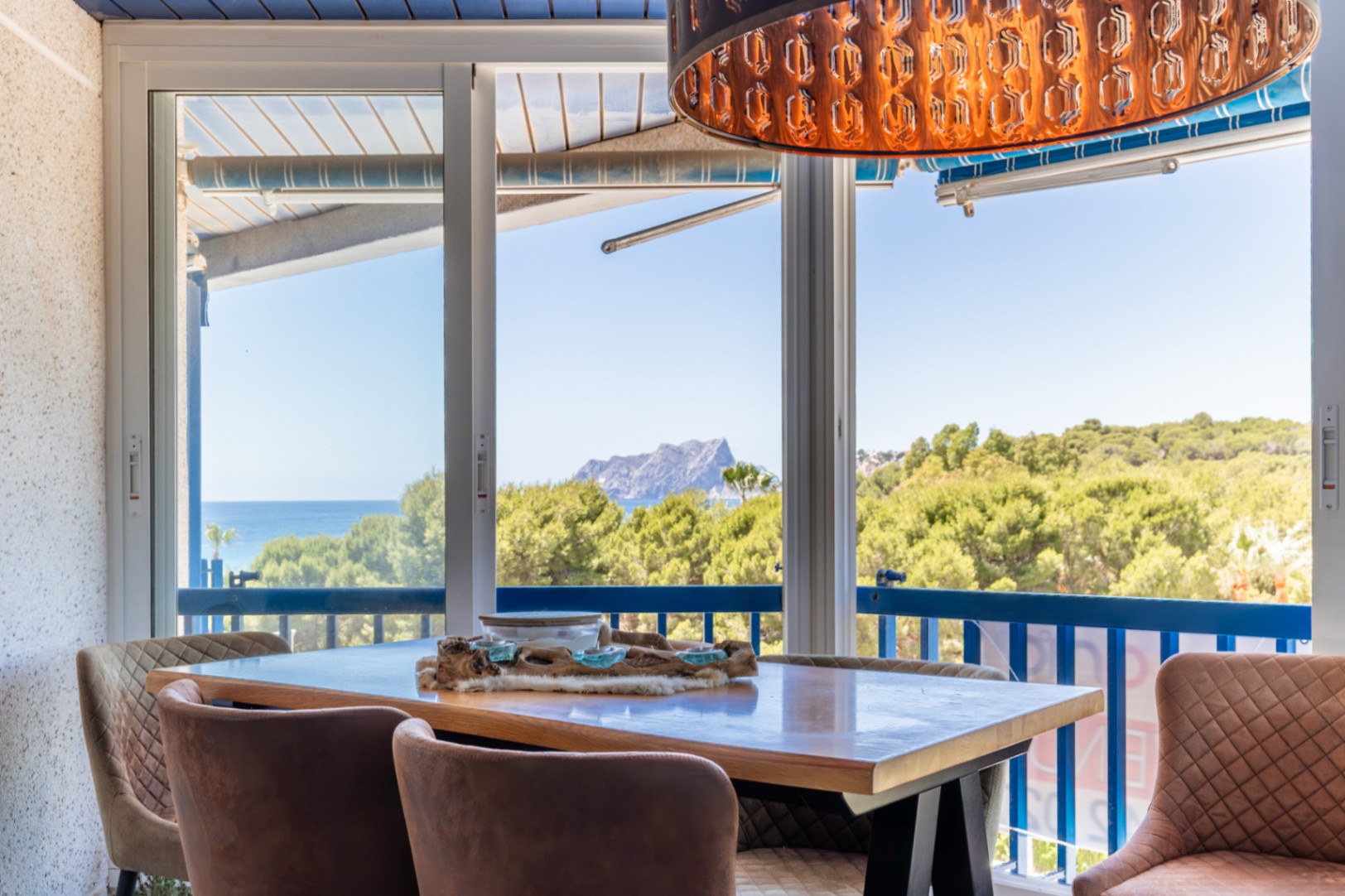 Espace repas lumineux avec vues panoramiques sur la mer, la montagne et la verdure via de larges baies vitrées. Une suspension décorative unique et des chaises rembourrées complètent l'ensemble.