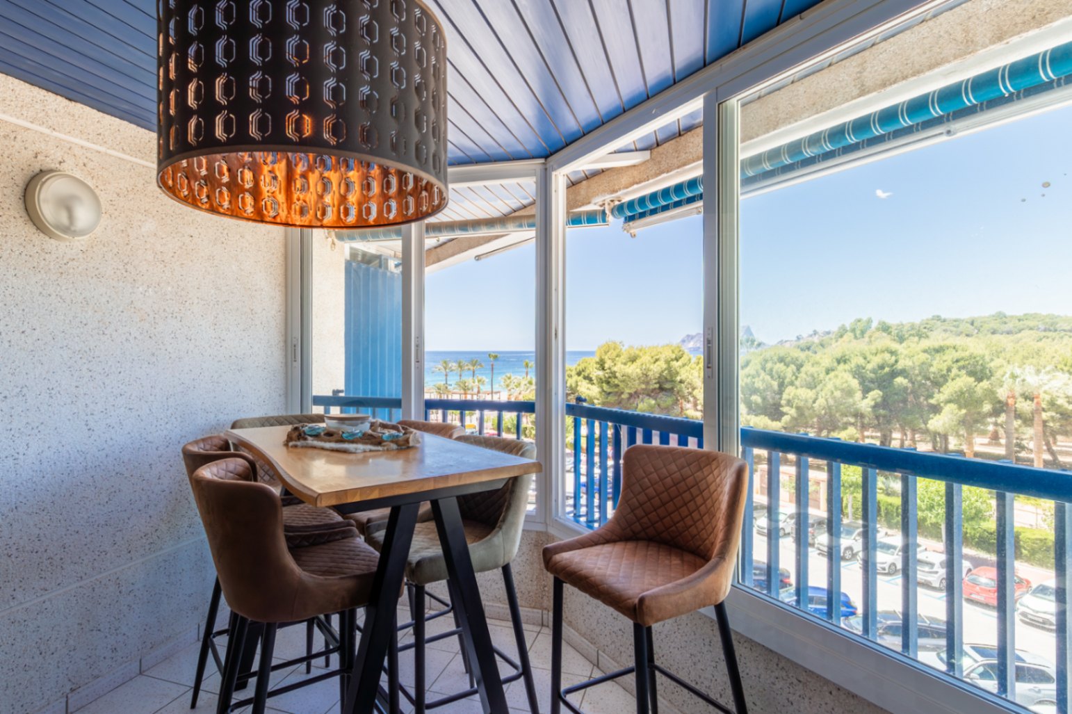 Espace repas vitré avec table haute en bois, tabourets rembourrés et suspension noire. Vues panoramiques sur mer, plage et pinède par les grandes fenêtres.