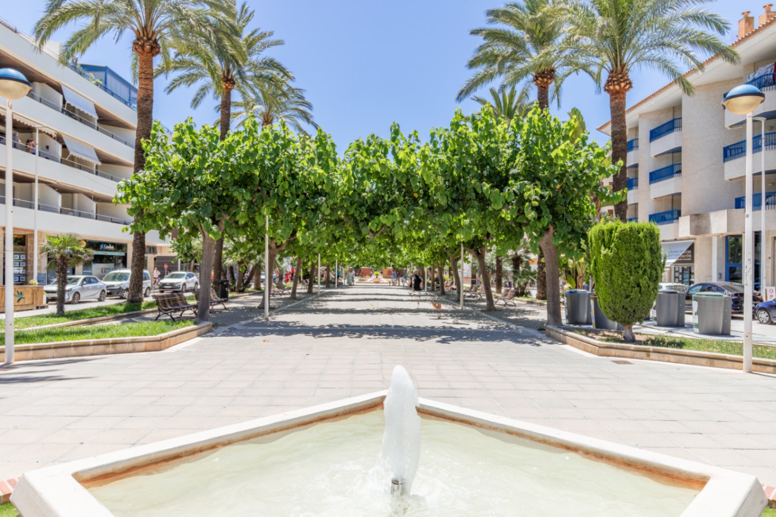 Large promenade piétonne à Moraira, avec une fontaine carrée au premier plan, ombragée par une canopée d'arbres verts et de grands palmiers, bordée d'immeubles.