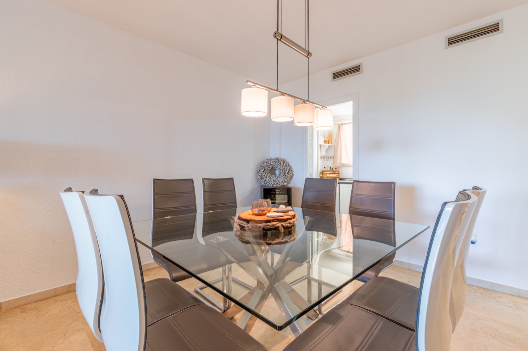 Salle à manger avec table carrée en verre et piètement métallique, entourée de chaises en cuir. Un luminaire suspendu moderne éclaire l'espace, relié à la cuisine.