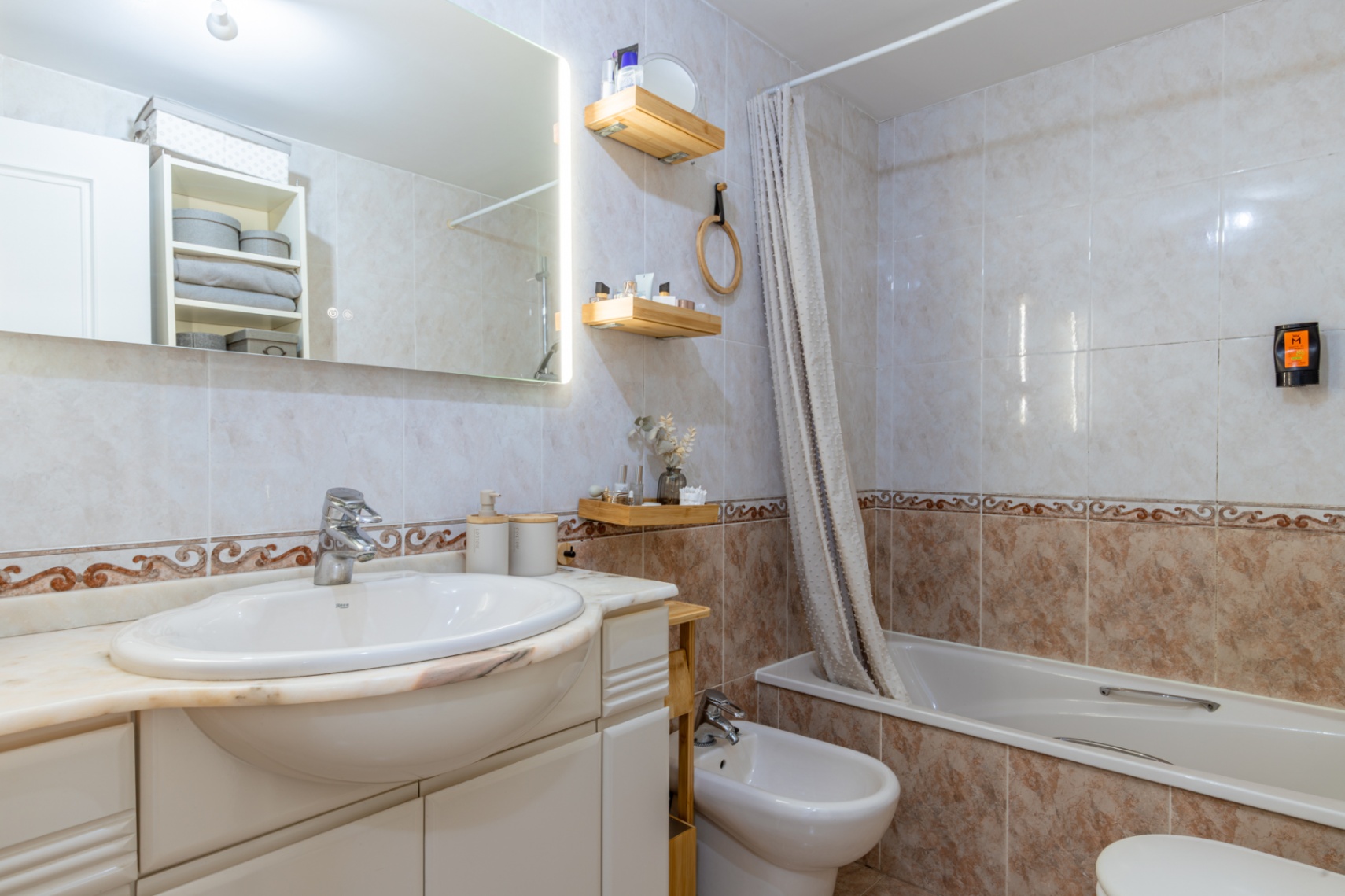 Salle de bain complète avec carrelage clair et frise décorative, lavabo intégré sur plan en marbre, miroir éclairé, baignoire avec rideau et bidet.