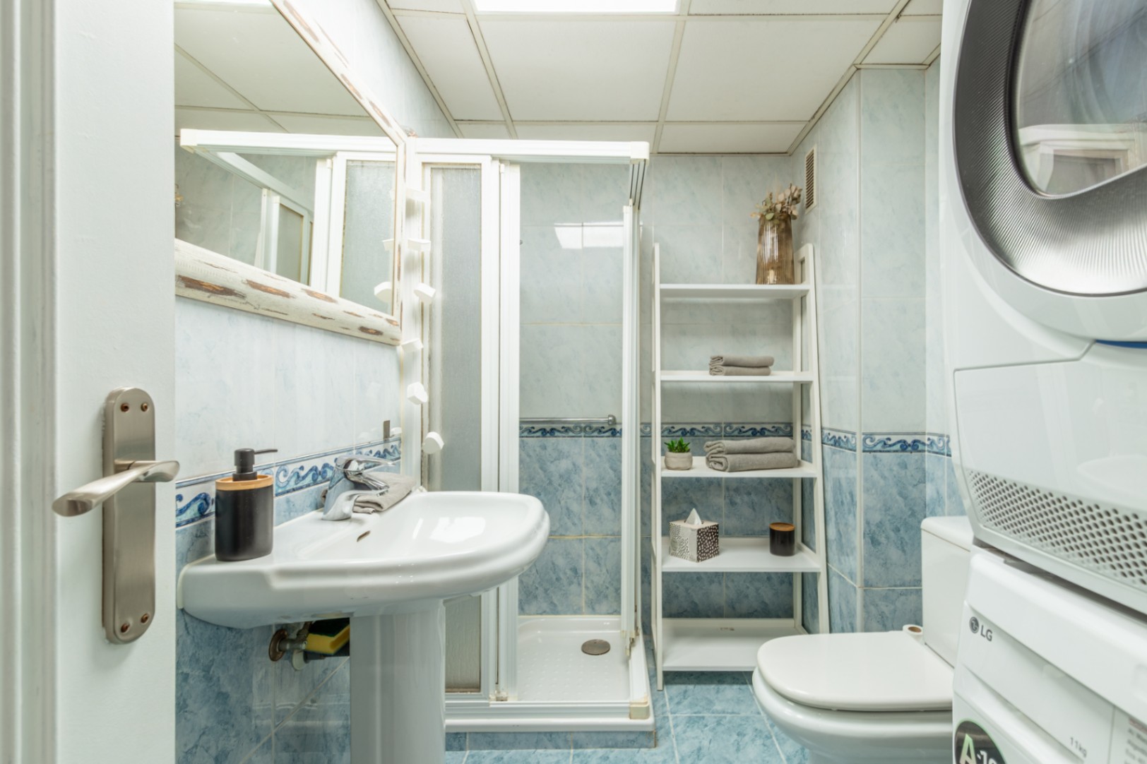 Salle de bain fonctionnelle avec carrelage bleu clair, lavabo sur pied, cabine de douche et appareils LG empilés. Une étagère échelle offre un rangement pratique.