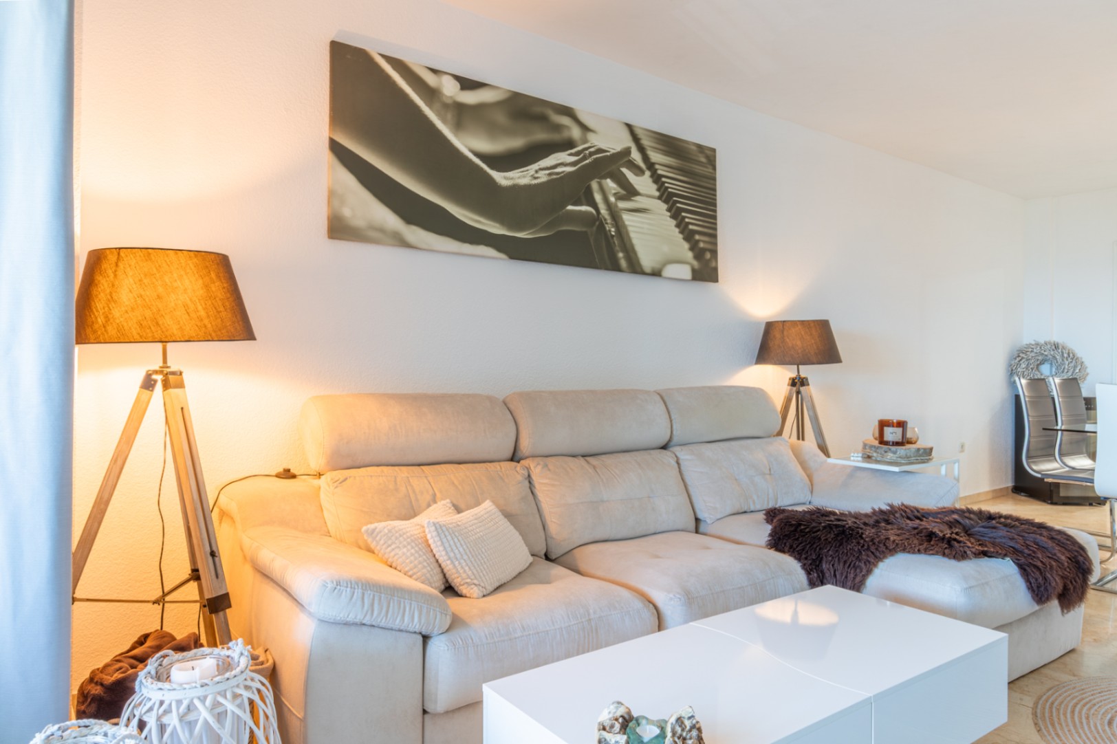 Salon avec canapé sectionnel clair, table basse blanche et deux lampadaires trépied en bois. Une grande œuvre d'art représentant un piano décore le mur.