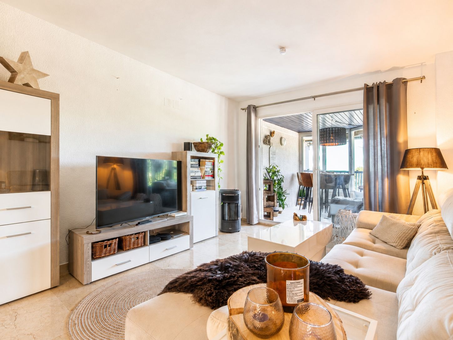 Salon lumineux avec un canapé d'angle clair, une table basse blanche brillante et un meuble TV. Des portes coulissantes en verre s'ouvrent sur une terrasse couverte.