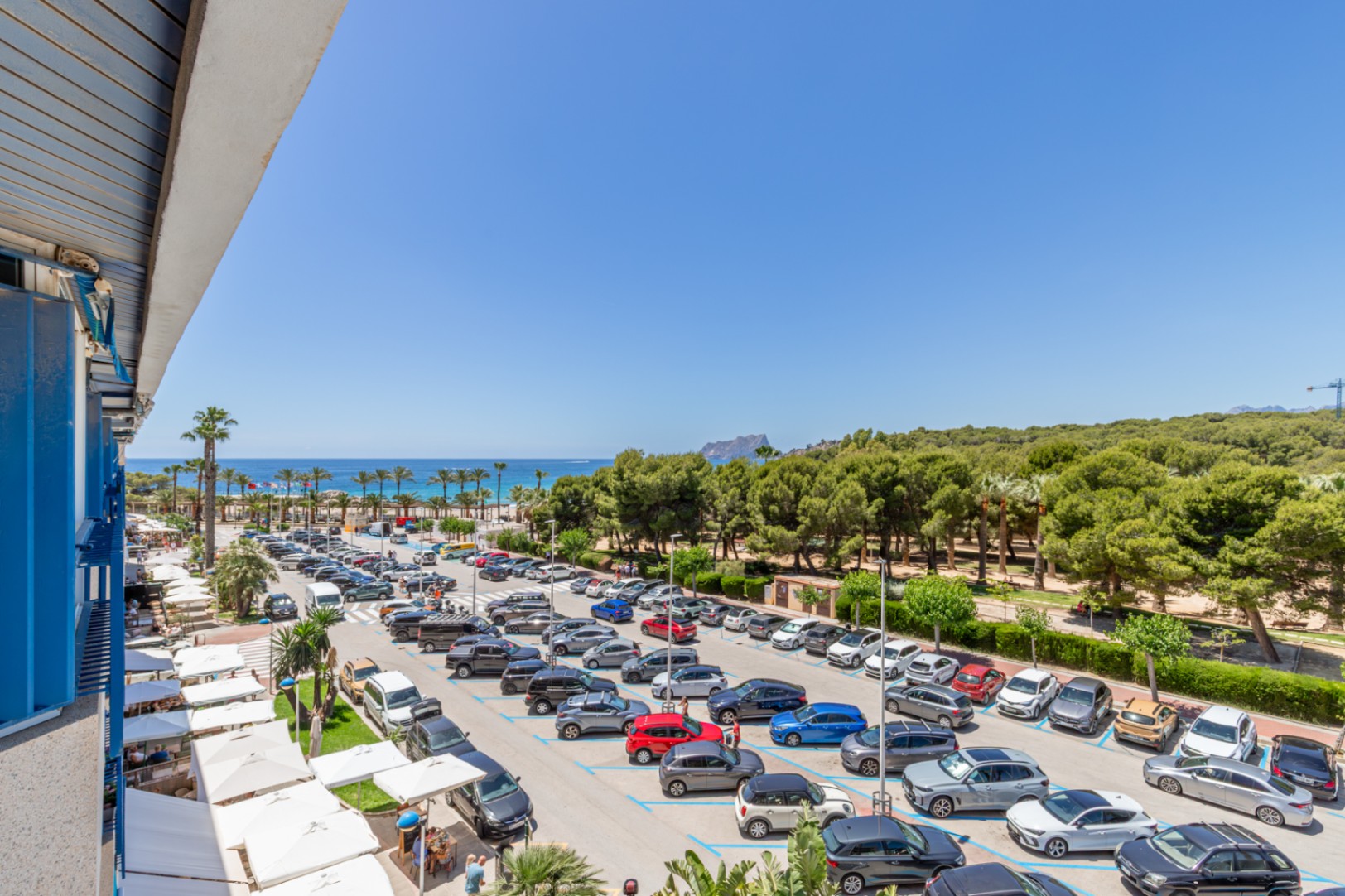 Vue panoramique depuis la terrasse, offrant la mer Méditerranée, une plage bordée de palmiers et le Peñón de Ifach. En contrebas, un parking et des auvents blancs.