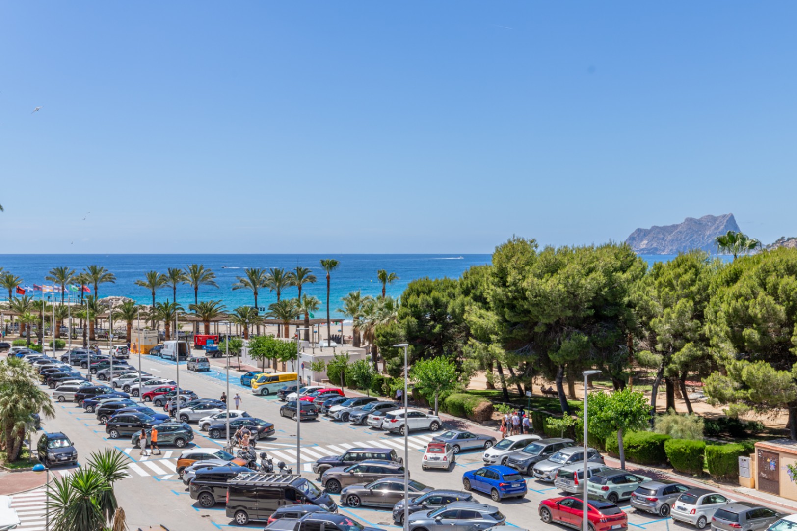 Vue panoramique sur la mer Méditerranée, avec des palmiers, une plage de sable et le Peñón de Ifach au loin, bordée par un parking animé et des pins.
