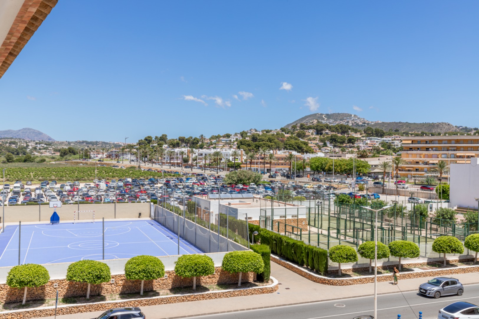Vue panoramique sur un terrain de sport bleu, plusieurs courts de padel, un vaste parking et le paysage urbain de Moraira avec des montagnes lointaines.