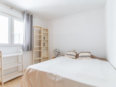 Chambre lumineuse avec lit double, étagères en bois clair et ventilateur. La fenêtre avec rideaux gris apporte une lumière naturelle abondante à l'espace aux murs blancs et au sol en marbre.