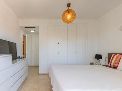 Chambre principale avec placards intégrés blancs, sol en marbre clair et luminaire suspendu sphérique. Comprend une commode avec TV et accès au couloir.