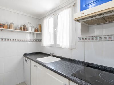 Cuisine indépendante avec plan de travail en granit foncé, évier blanc et plaque à induction. Murs carrelés blancs avec frise décorative et fenêtre apportant lumière naturelle.