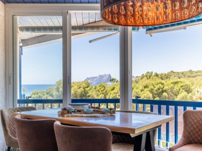 Espace repas lumineux avec vues panoramiques sur la mer, la montagne et la verdure via de larges baies vitrées. Une suspension décorative unique et des chaises rembourrées complètent l'ensemble.