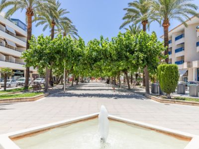 Large promenade piétonne à Moraira, avec une fontaine carrée au premier plan, ombragée par une canopée d'arbres verts et de grands palmiers, bordée d'immeubles.