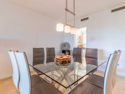 Salle à manger avec table carrée en verre et piètement métallique, entourée de chaises en cuir. Un luminaire suspendu moderne éclaire l'espace, relié à la cuisine.