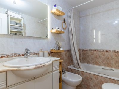 Salle de bain complète avec carrelage clair et frise décorative, lavabo intégré sur plan en marbre, miroir éclairé, baignoire avec rideau et bidet.